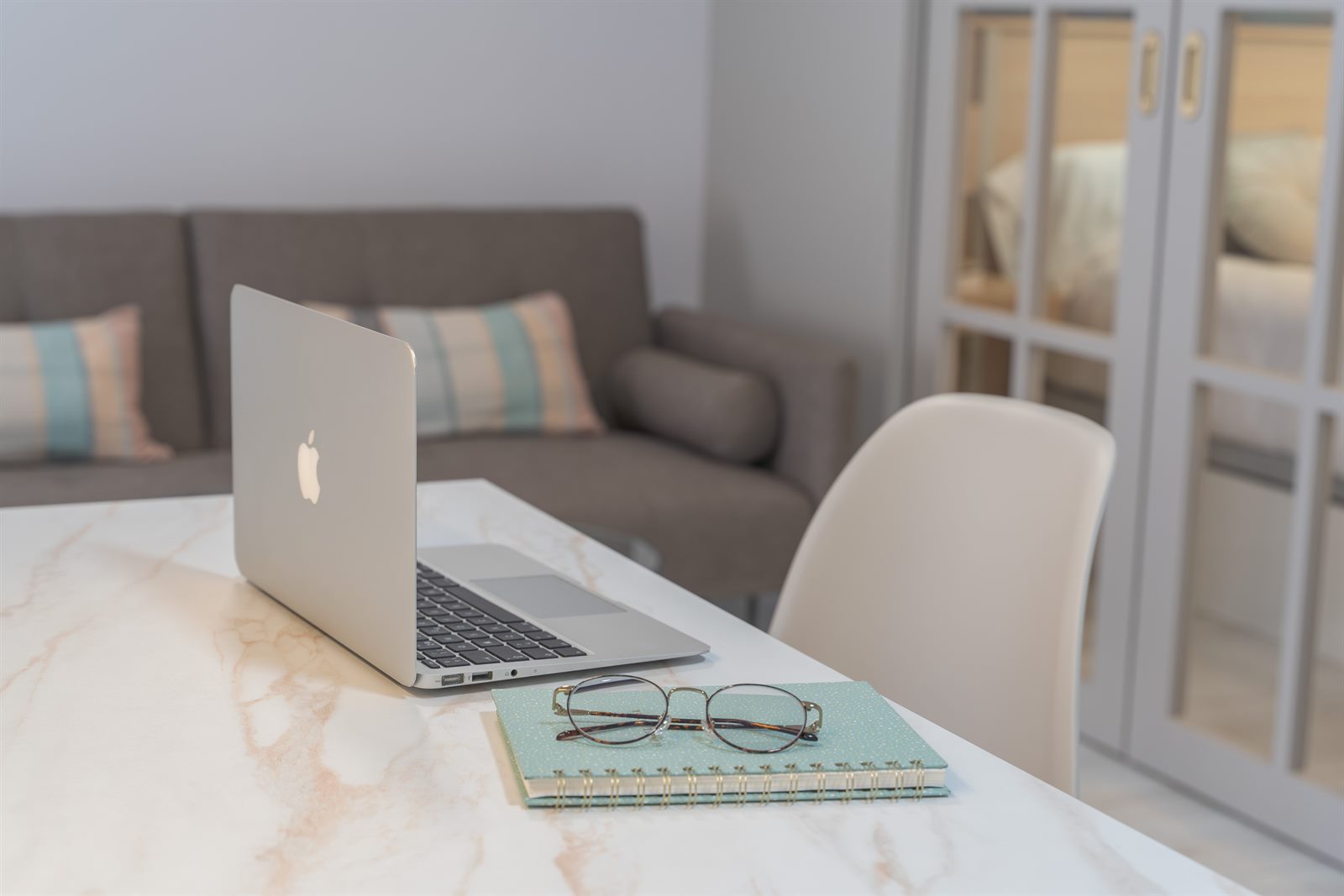 Portátil abierto sobre una mesa de mármol con gafas y una libreta, junto a un sofá gris